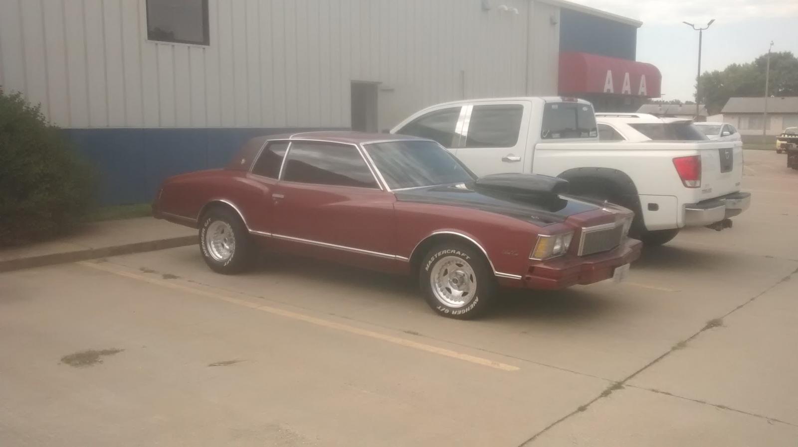 A classic maroon Chevrolet Monte Carlo and pickups in the Car Guys lot